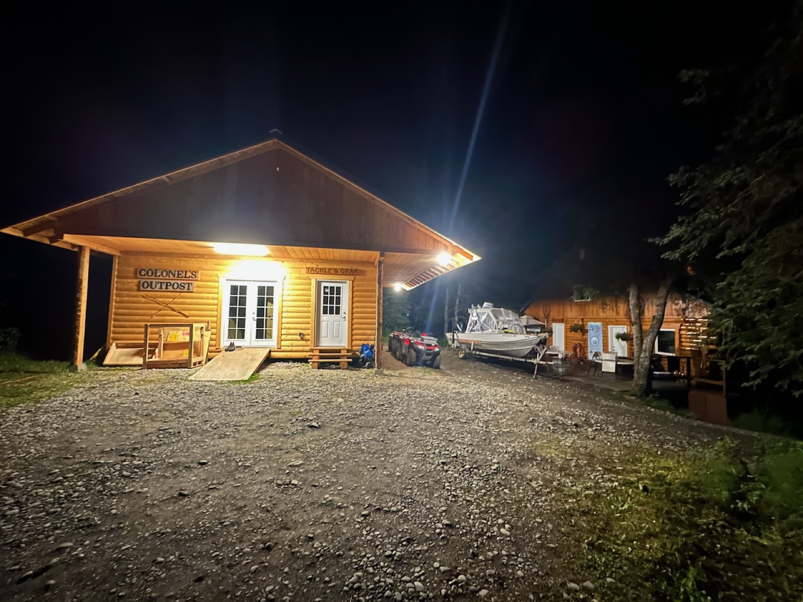 Colonel's Outpost log cabin at night — signage lit, boat parked under the overhang, lodge building behind