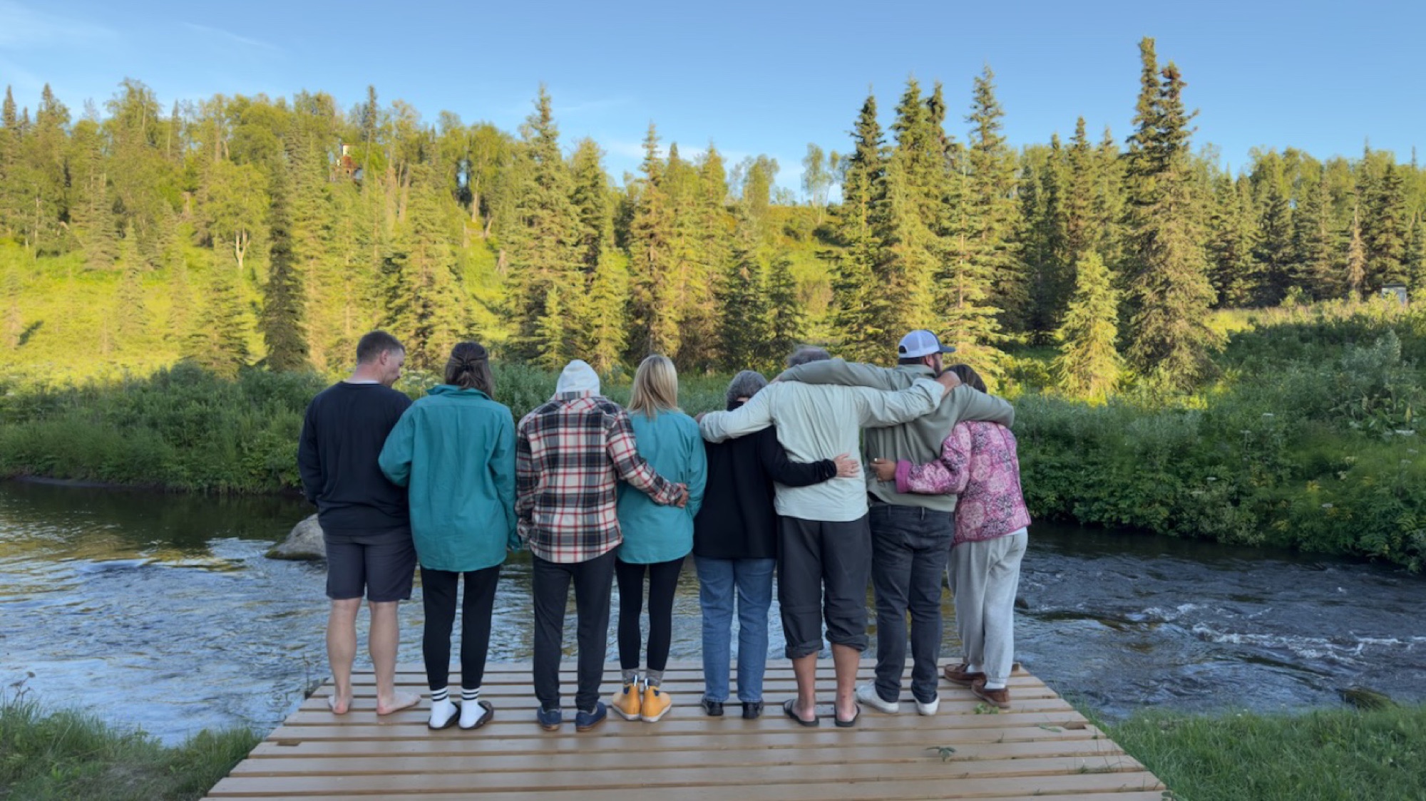 Eight people standing arm-in-arm on the deck overlooking the river through spruce, evening light