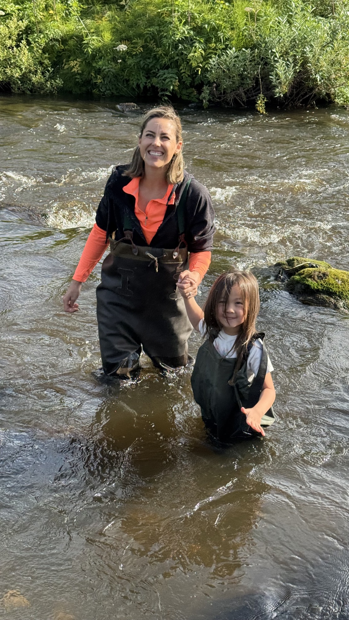 Janelle and Juno in chest waders, both grinning, waist-deep in the river
