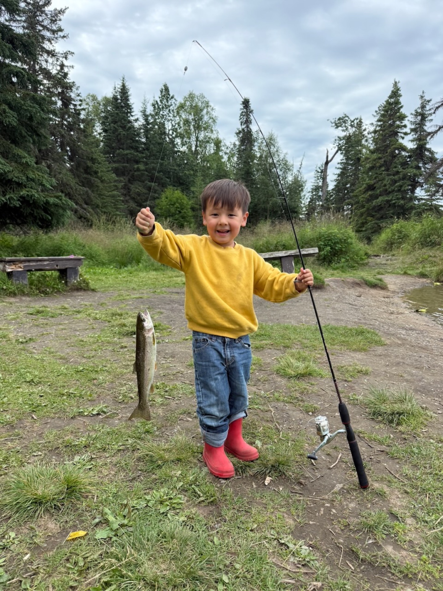 Juno in a yellow sweater and red boots grinning, holding a rod with his first salmon