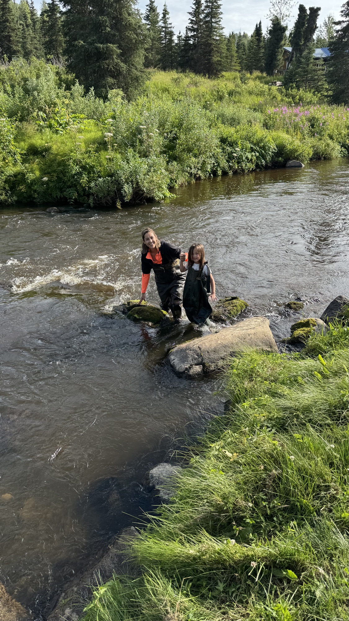Janelle and Juno in chest waders, stepping across moss-covered rocks in the river, lodge visible behind them