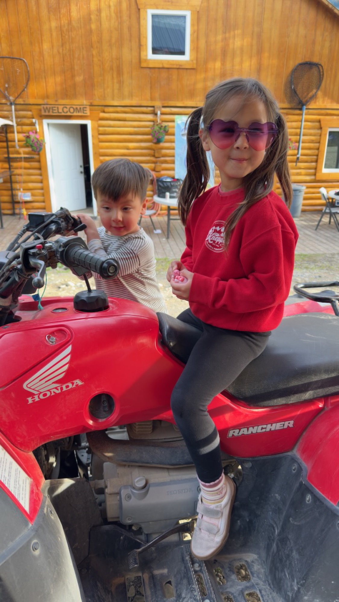 Two kids sitting on a red Honda Rancher ATV in front of the lodge