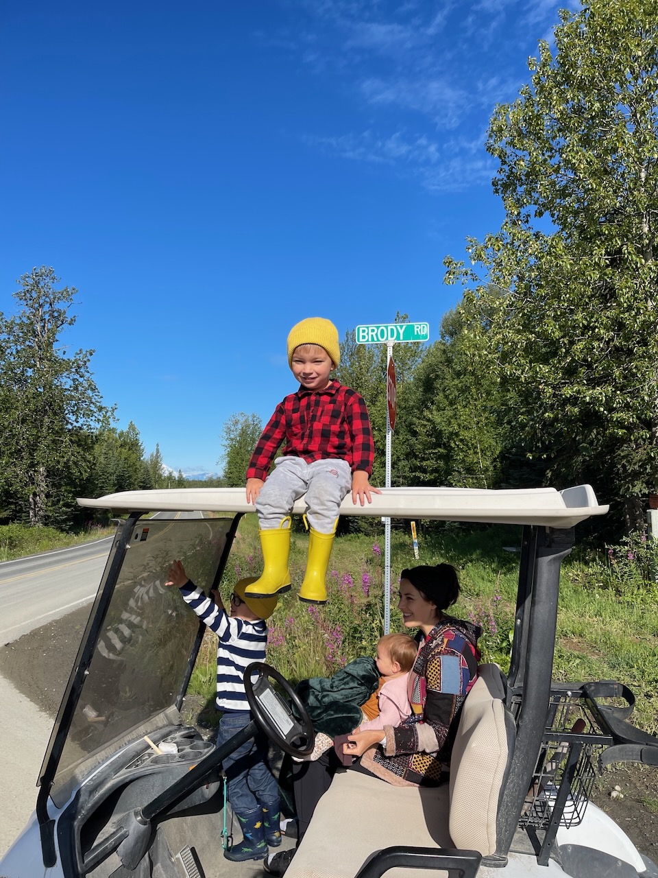 Kid in yellow beanie and red flannel sitting on top of the golf cart at the Brody Road sign