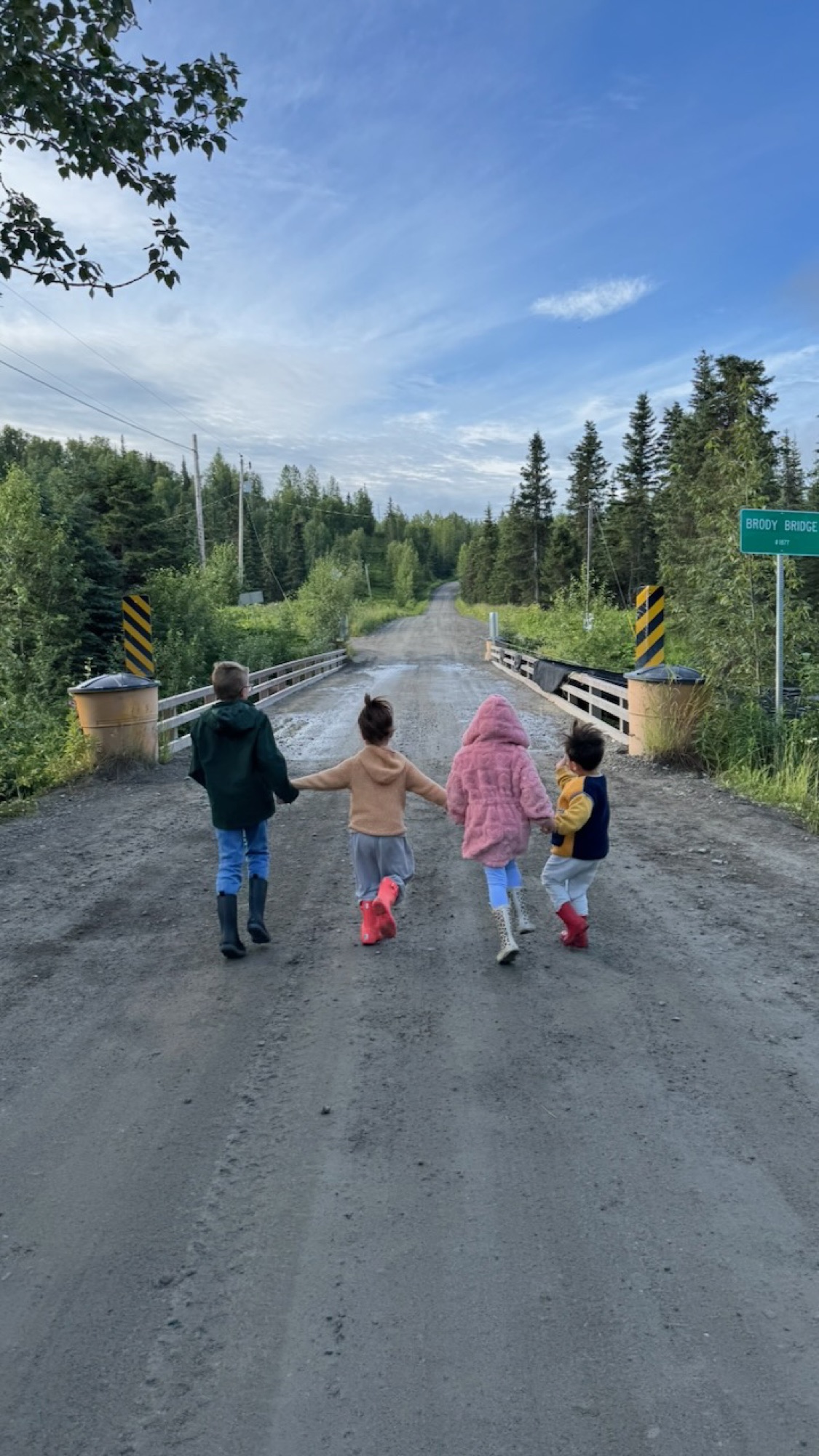 Four kids in jackets and rubber boots holding hands, walking down the dirt road toward Brody Bridge