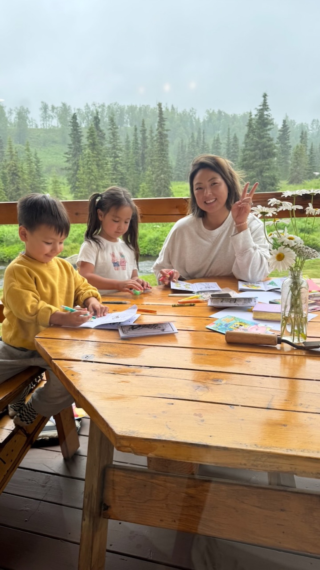 Janelle and her two kids coloring at the porch table, misty river and spruce behind